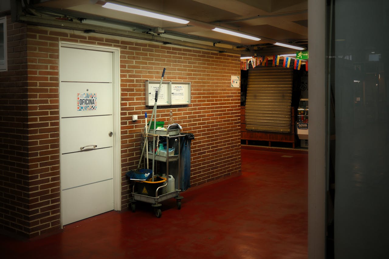 about-01 A brightly lit industrial hallway with a cleaning cart and supplies near an office door.